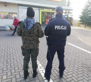 Na zdjęciu policjant i żołnierz stojący przed wejściem do marketu.
